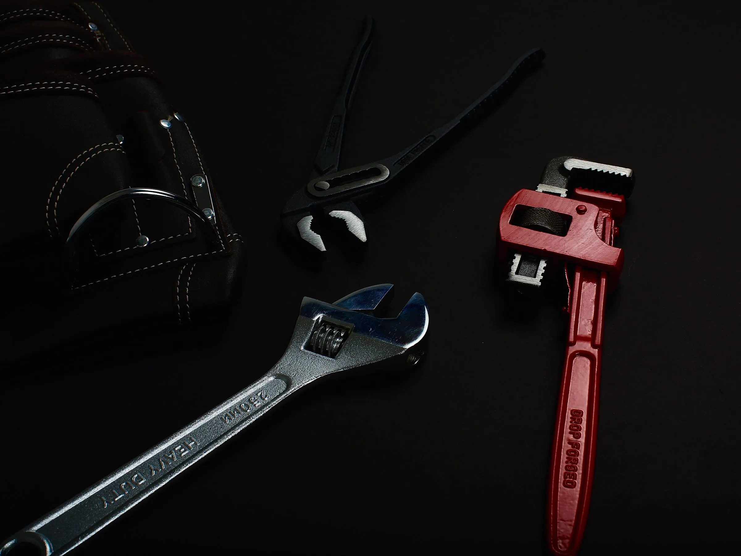 A pipe wrench, adjustable wrench and pliers laid out on a dark workbench